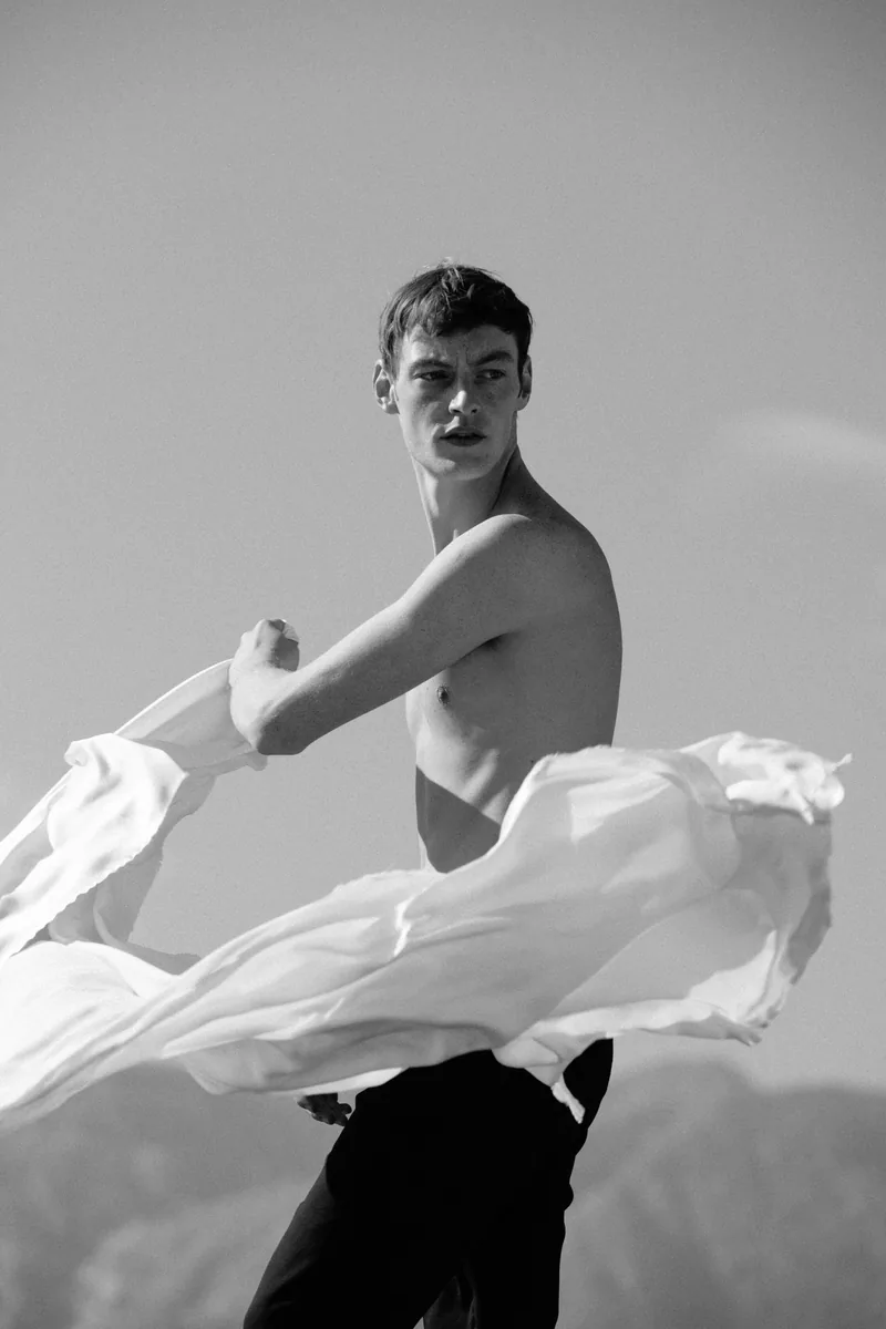 Black-and-white photograph of a model sweeping white fabric around his waist outdoors.