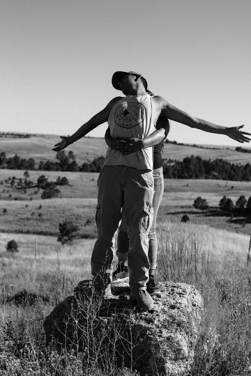 Black-and-white photograph of a person standing on a rock with arms outstretched over open countryside.
