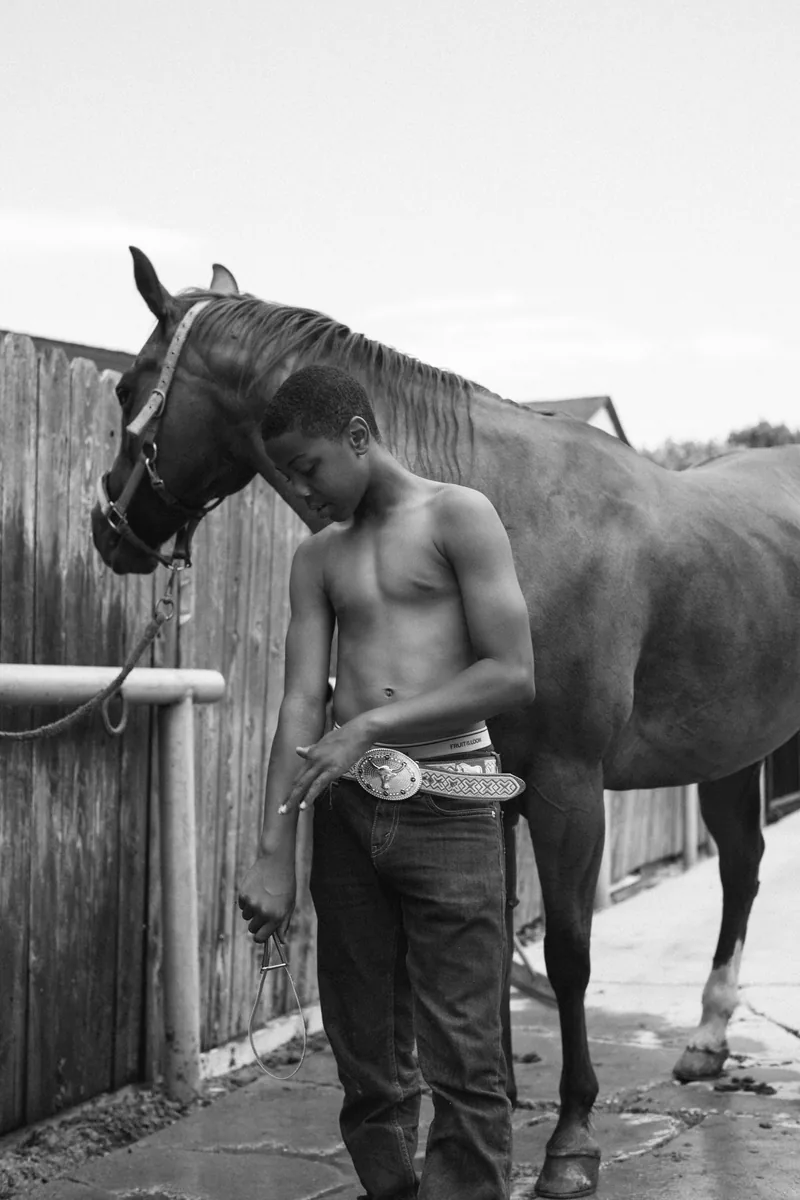 Black-and-white photograph of a young person looking down beside a horse at a wooden fence.
