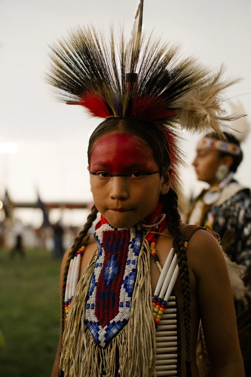 A child wearing beaded and feathered regalia with red face paint.