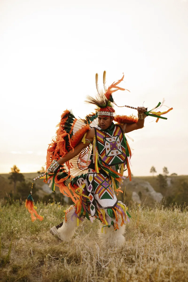 A dancer in colorful beaded and feathered regalia moving through tall grass.