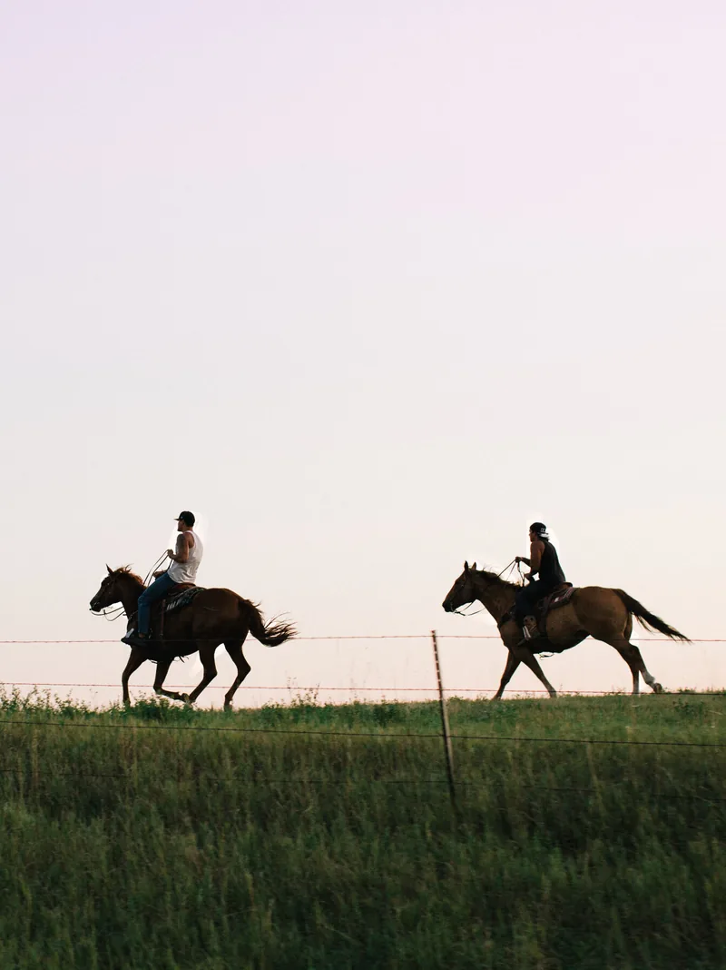 Two riders crossing a grassy ridge on horseback beneath a pale sky.