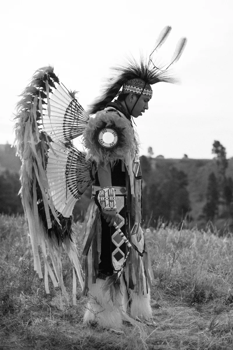 Black-and-white portrait of a person in feathered regalia standing in tall grass.
