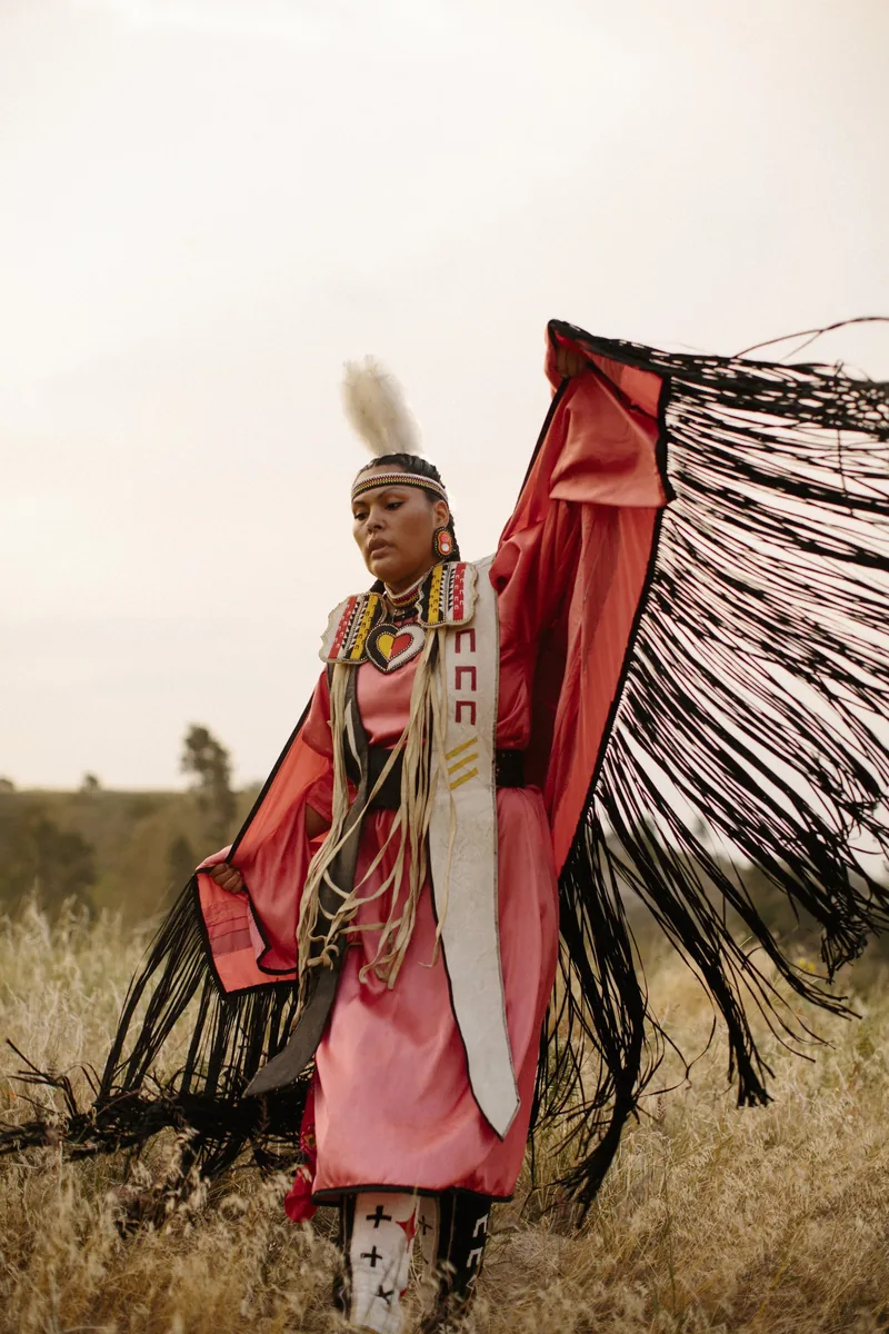 A person in pink regalia holding a fringed shawl out to one side in a field.