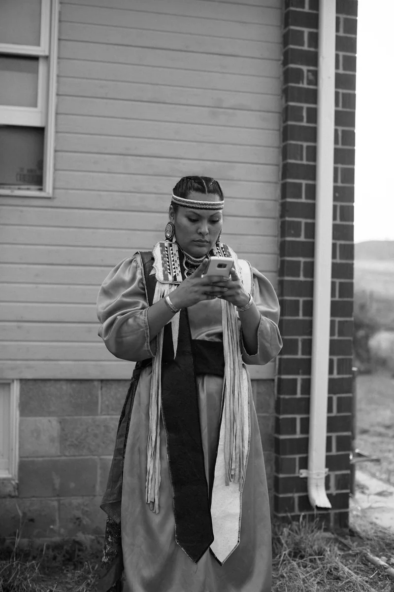 Black-and-white photograph of a person in regalia looking down at their hands outside a house.