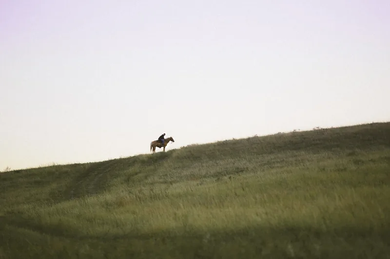A distant horse rider crossing a green hillside beneath a pale sky.