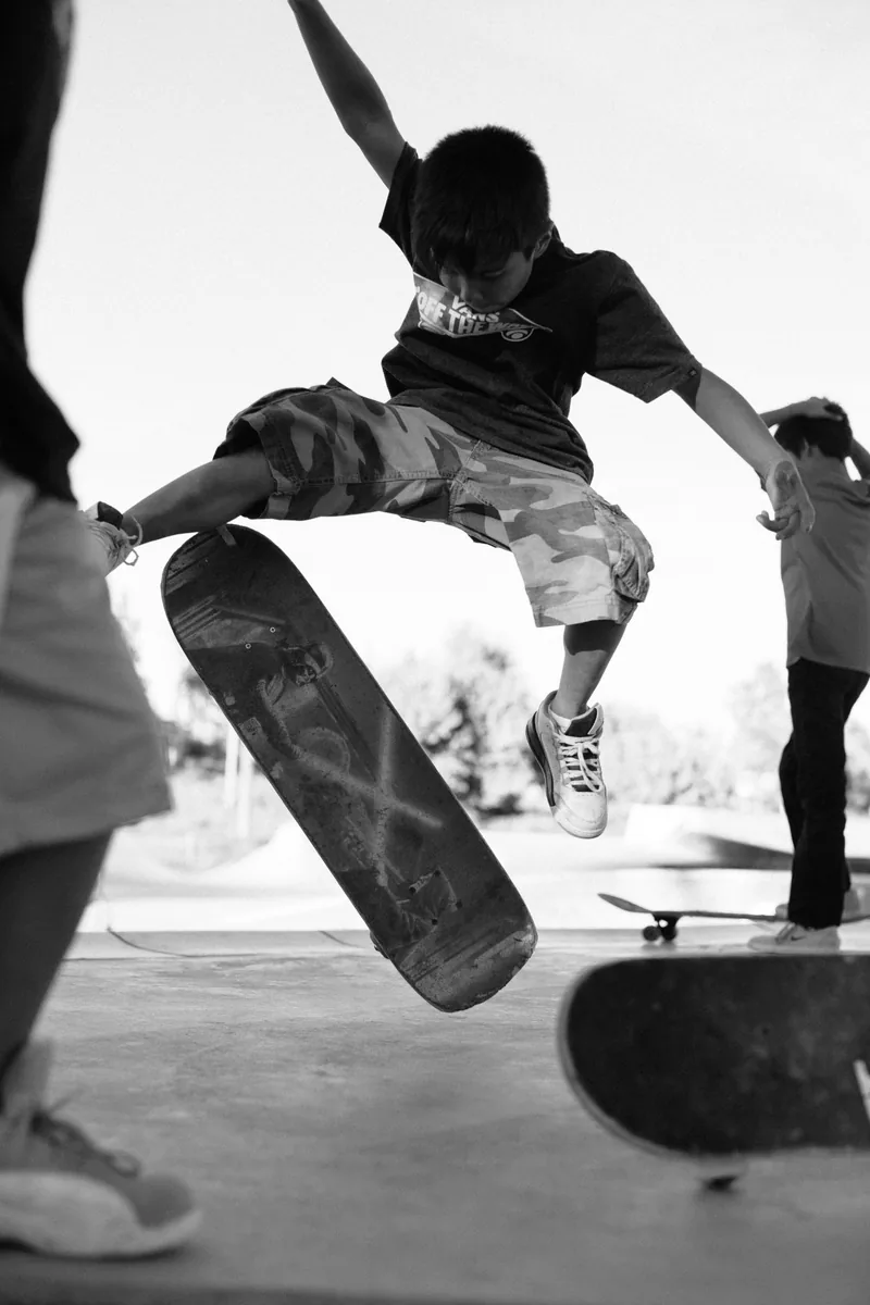 Black-and-white action photograph of a child jumping above a skateboard at a skate park.