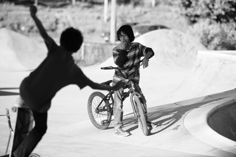 A child on a bicycle facing another child in a concrete skate bowl, in black and white.