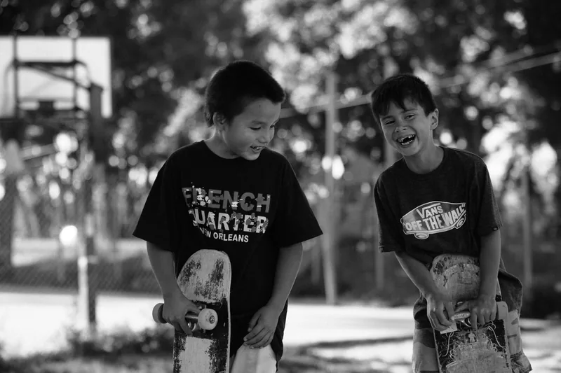Two children laughing together while holding skateboards, photographed in black and white.