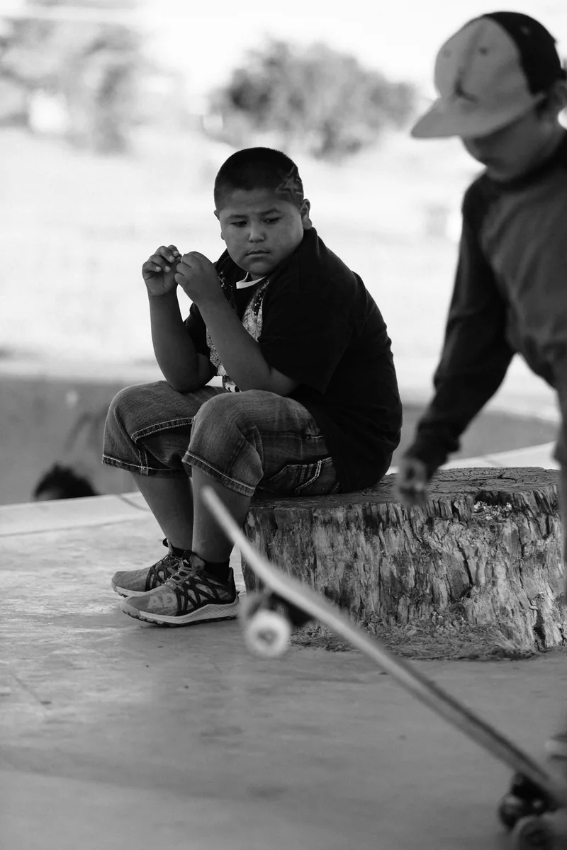 A child sitting at the edge of a skate park beside a skateboard, in black and white.