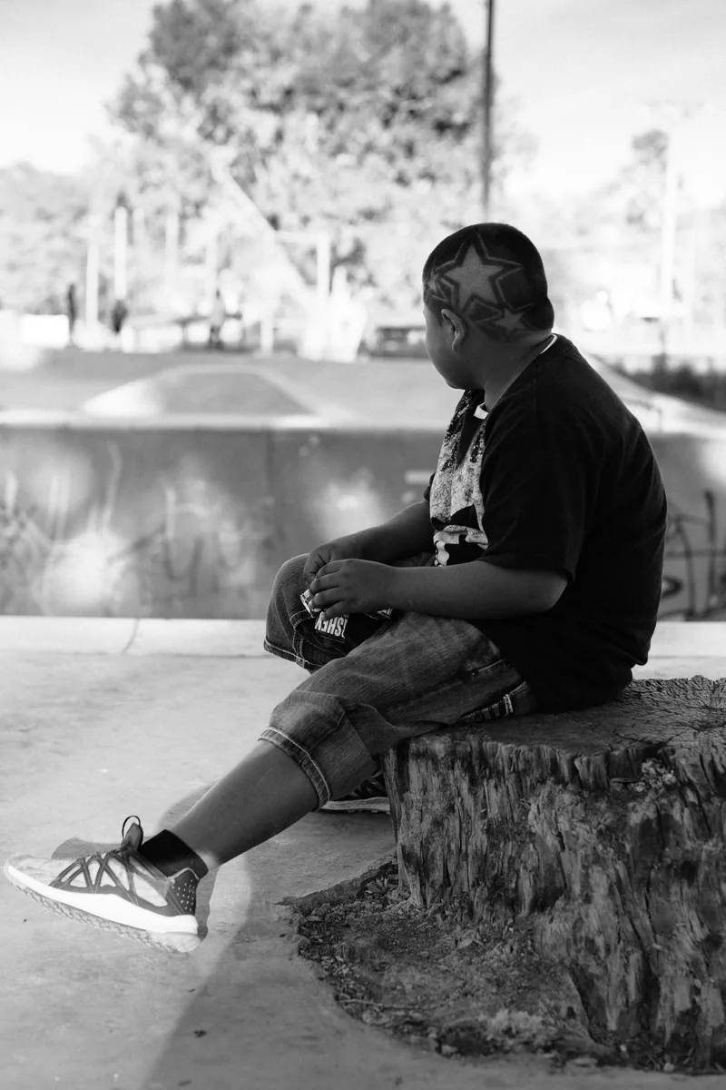 Black-and-white portrait of a child seated on a low ledge at a skate park.